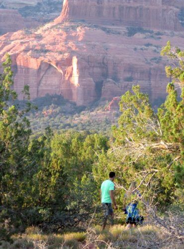 2 people walking under trees with red cliffs of Sedona in the background. This symbolizes attentiveness in nature practice to help participants discover nature as a portal to spiritual awareness.
