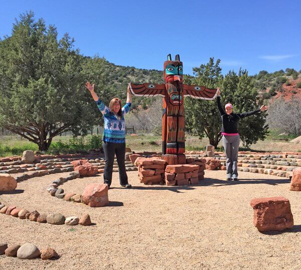 2 people arms lifted toward sky are standing next to a totem pole in center of concentric rings medicine wheel. This symbolizes the power animal activation ceremony we do during our Power Animal connections Sedona workshop.